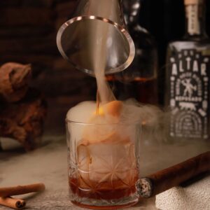 Mariposas Ancestral Smoked Old Fashioned with an orange slice, cinnamon, and aromatic smoke in a patterned glass.