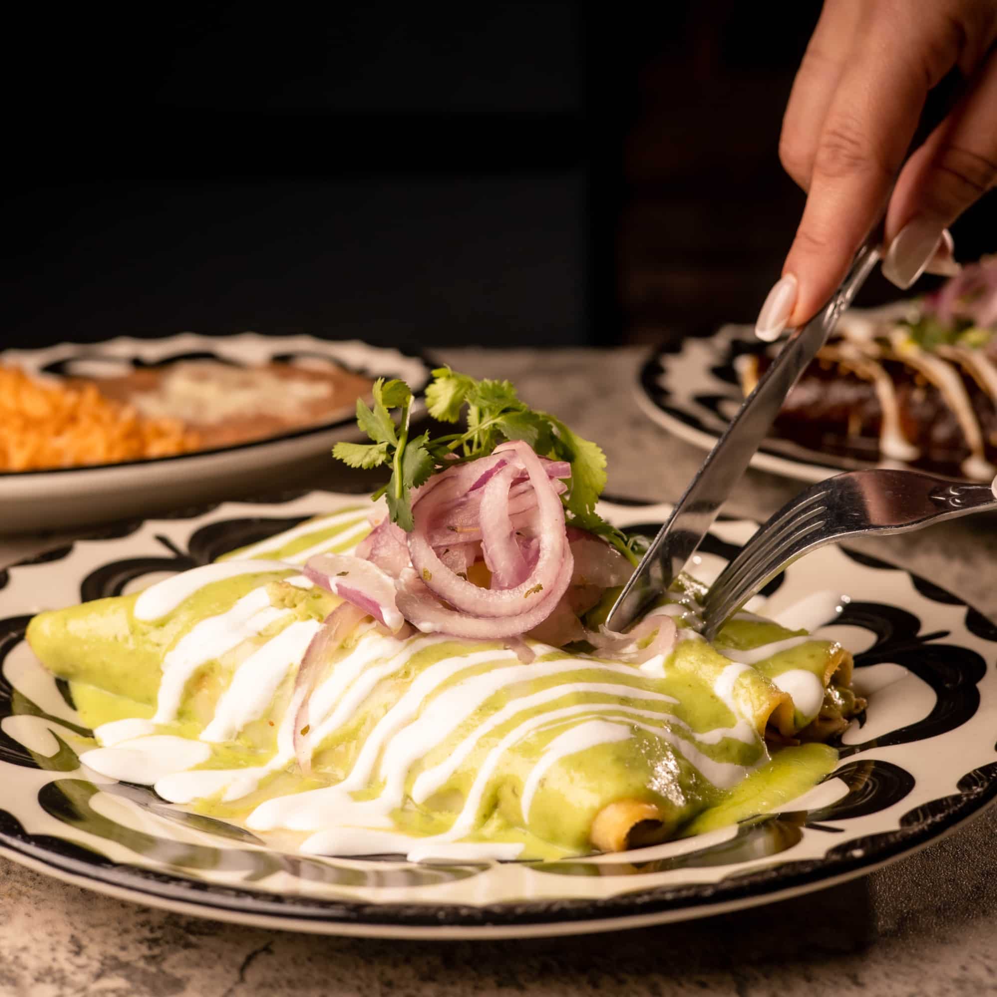 kulturas_enchiladas_verdes A person using a fork and knife to slice into enchiladas covered in green sauce and white crema. The enchiladas are topped with cilantro and thinly sliced red onions and placed on a decorative black and white plate. Rice and beans are blurred in the background.