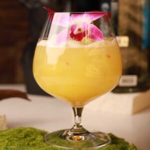 Mariposas Lindo Amanecer, a golden tropical cocktail with a frothy top and vibrant orchid garnish.