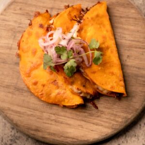 Two golden Mariposas quesabirria tacos filled with braised beef and melted cheese, topped with red onion and cilantro.
