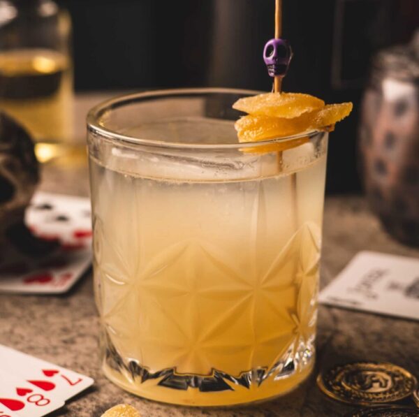 A cocktail in a textured glass filled with a pale yellow drink is garnished with candied ginger on a stick topped with a small purple skull. Playing cards, a skull decoration, and a gold coin are scattered on the surface around the glass, creating a mysterious setting.