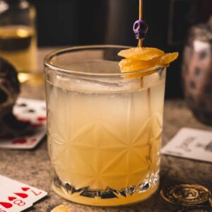 Mariposas Raicillina, a pale golden cocktail in a textured glass with a candied ginger garnish.