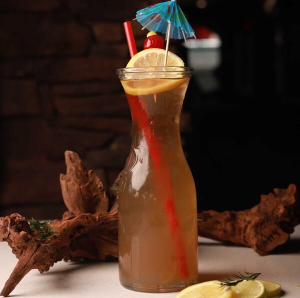kulturas_riverside_water A tall glass of iced tea is garnished with a lemon slice, cherry, blue cocktail umbrella, and red straw. It sits on a table with wooden decor in the background, with lemon slices nearby.