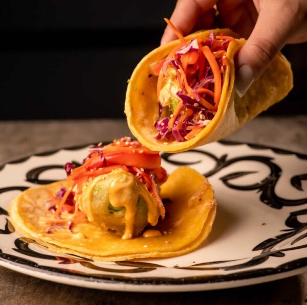 A person is holding a taco above a plate. The taco has a yellow shell filled with a creamy sauce, shredded purple and orange cabbage, and carrots. The plate beneath holds another taco with similar fillings, featuring a decorative black and white pattern.