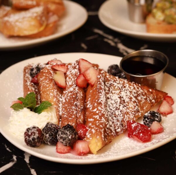kulturas_french_toast A plate of French toast dusted with powdered sugar, topped with sliced strawberries and blackberries. A dollop of whipped cream and a sprig of mint are on the side. A metal cup of syrup accompanies the dish. Another plate is partially visible in the background on a dark surface.