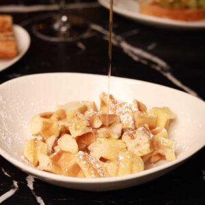 Mariposas bite-size Belgian waffle pieces dusted with powdered sugar as warm syrup is poured over them.