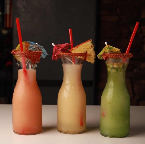 Three tall glass bottles filled with colorful drinks—pink, yellow, and green—are garnished with fruit slices, small umbrellas, and chili powder on the rims. Each has a red straw. The bottles are arranged on a white surface with a dark background.