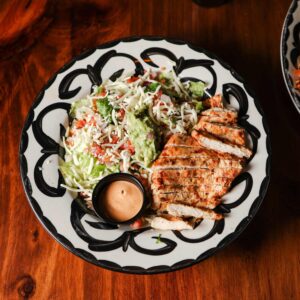Mariposas Burrito Bowl with grilled chicken, romaine, tomato, shredded cheese, guacamole, and creamy dressing on a patterned plate.
