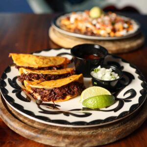 Three Mariposas quesabirria tacos filled with shredded beef, served with lime, onion, cilantro, and consommé.