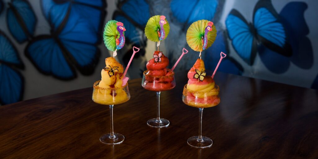 Three colorful soft serve margaritas with playful garnishes against the blue butterfly mural at Mariposas.